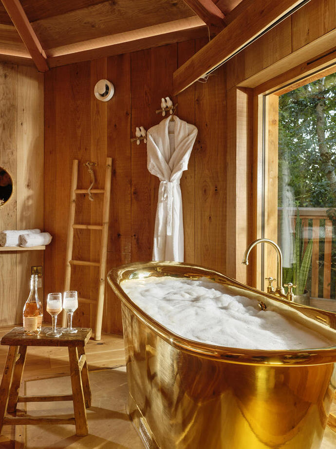 The interior of a wooden lodge, with a gold roll-top bath by the window at Sleepy Owl Devon.