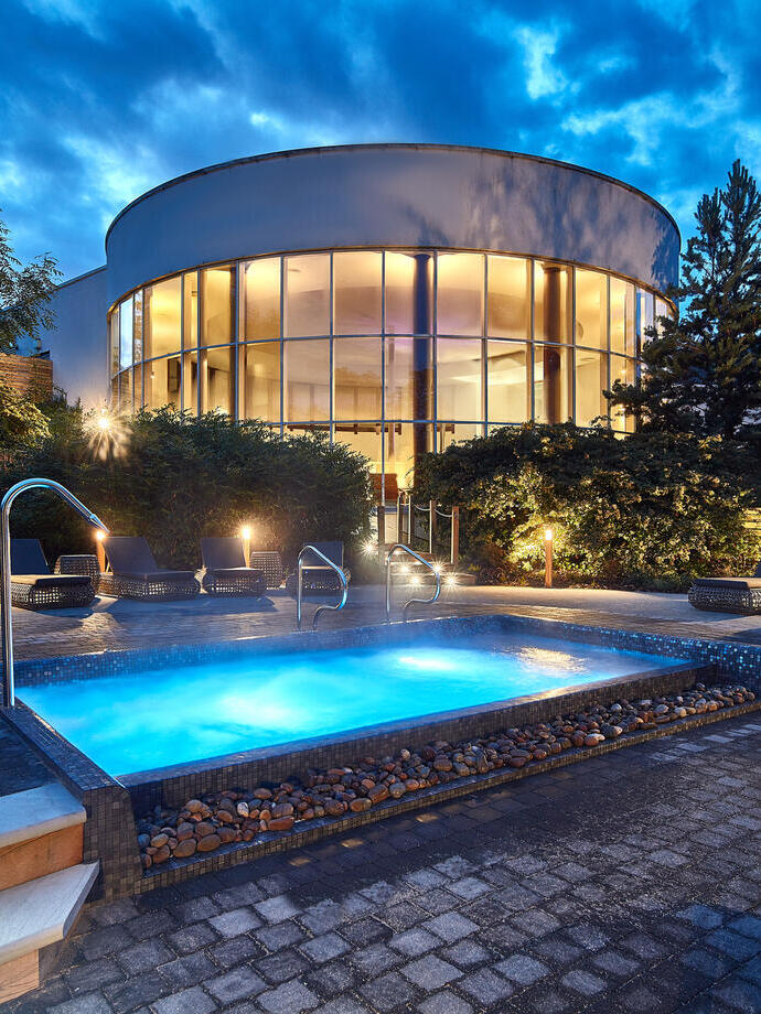 The outdoor spa area at dusk with a lit outdoor spa pool surrounded by loungers and the glass fronted circular spa building in the background. 