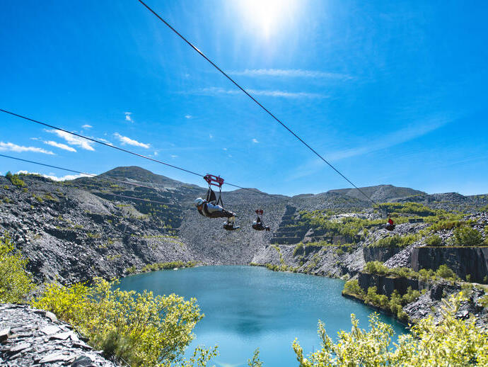 People ziplining over a blue lake in a rocky quarry landscape under a clear sunny sky, surrounded by hills and scattered greenery.
