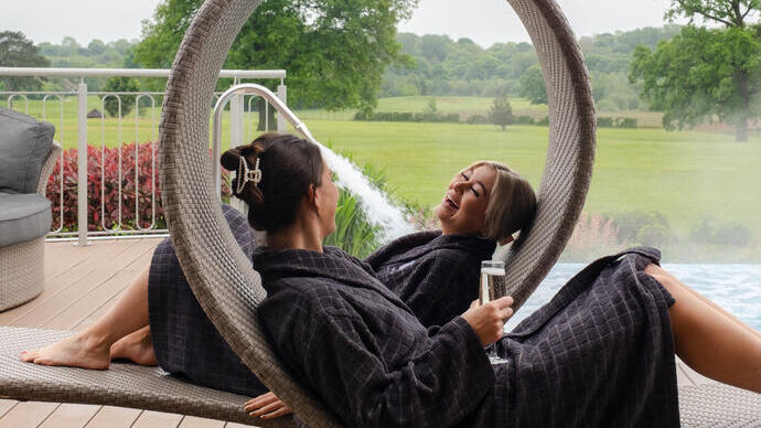 Two women in robes lay on loungers with a garden beyond