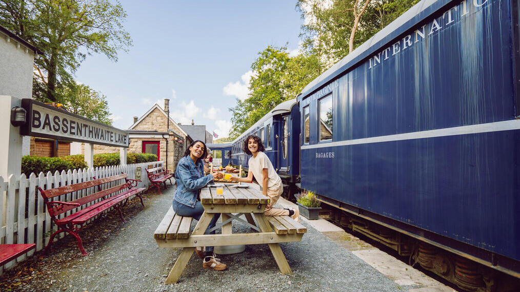 Two people dining at a picnic table on a historic train station platform with a blue train and Bassenthwaite Lake station sign visible.