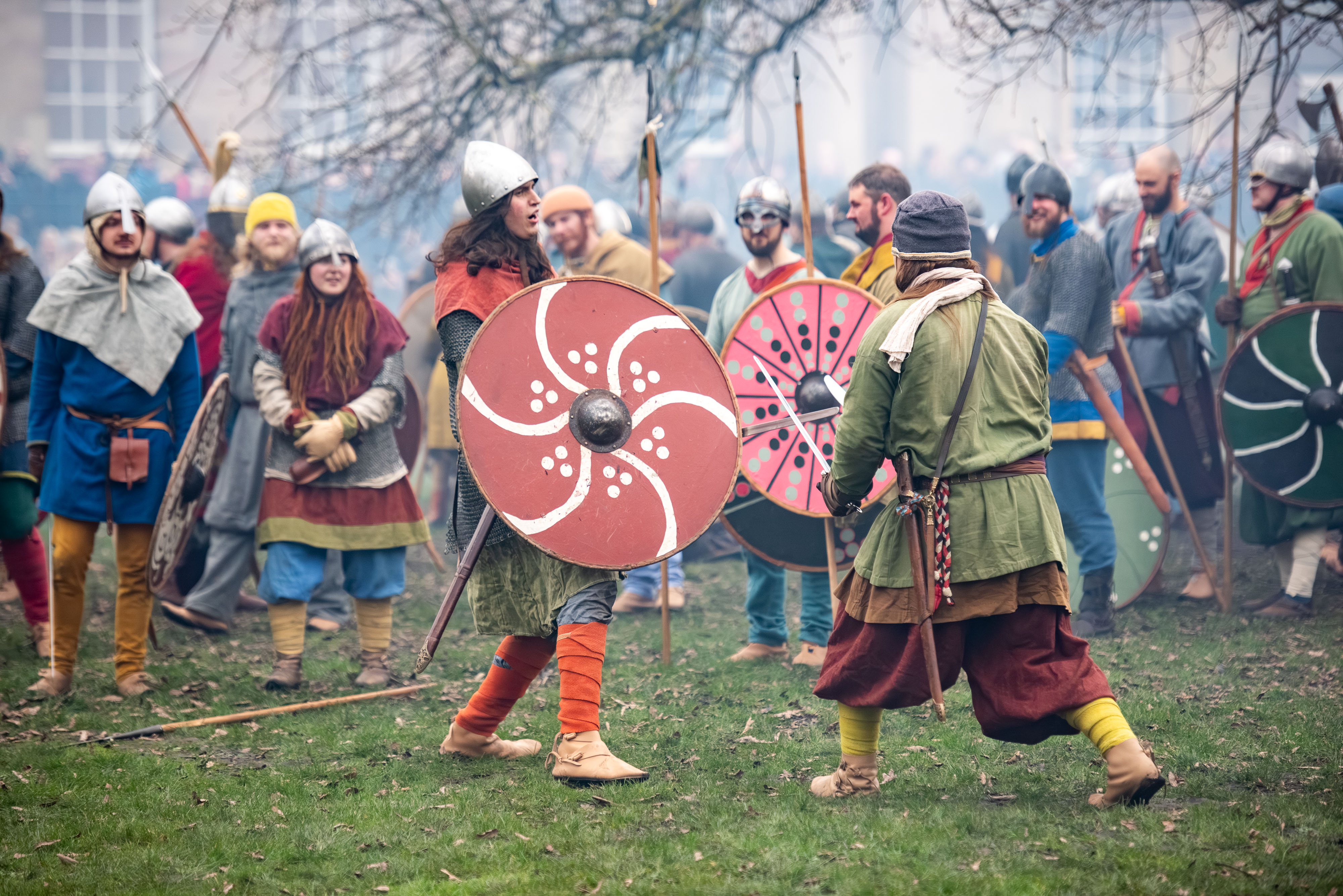 People in historical Viking attire with shields and helmets reenact a battle outdoors, with spectators and trees in the background.