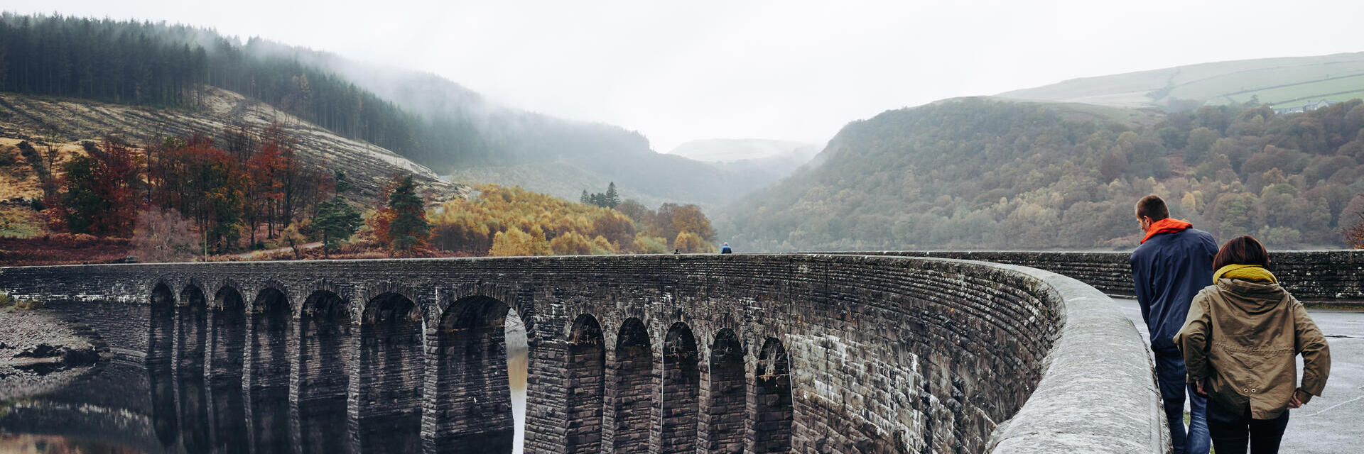 Two people walking on an arched bridge spanning a lake, with a valley in the background