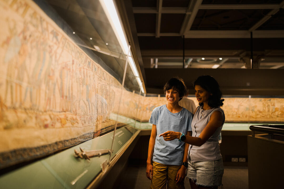 Two people observe an embroidered tapestry displayed in a museum exhibit under glass.
