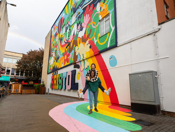 Two people playfully smiling and posing in front of a colourful mural.