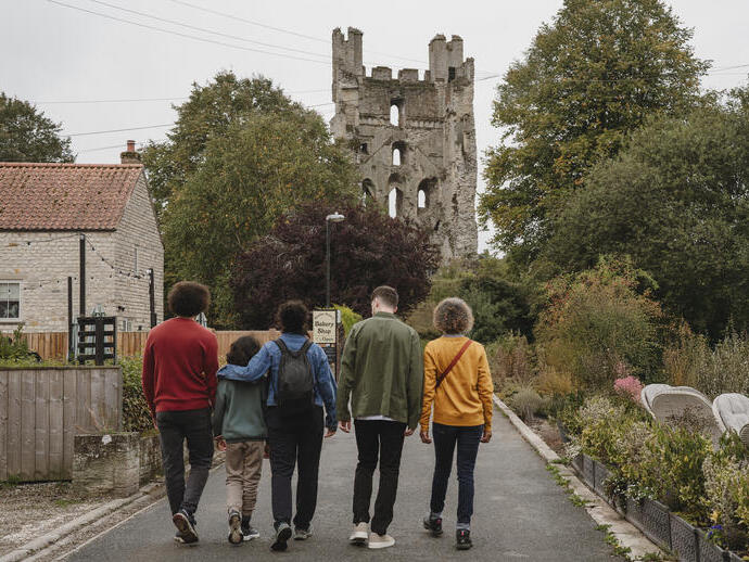 Five people walk towards a stone tower ruin, with a house and garden beside the road. Trees and greenery surround the area.