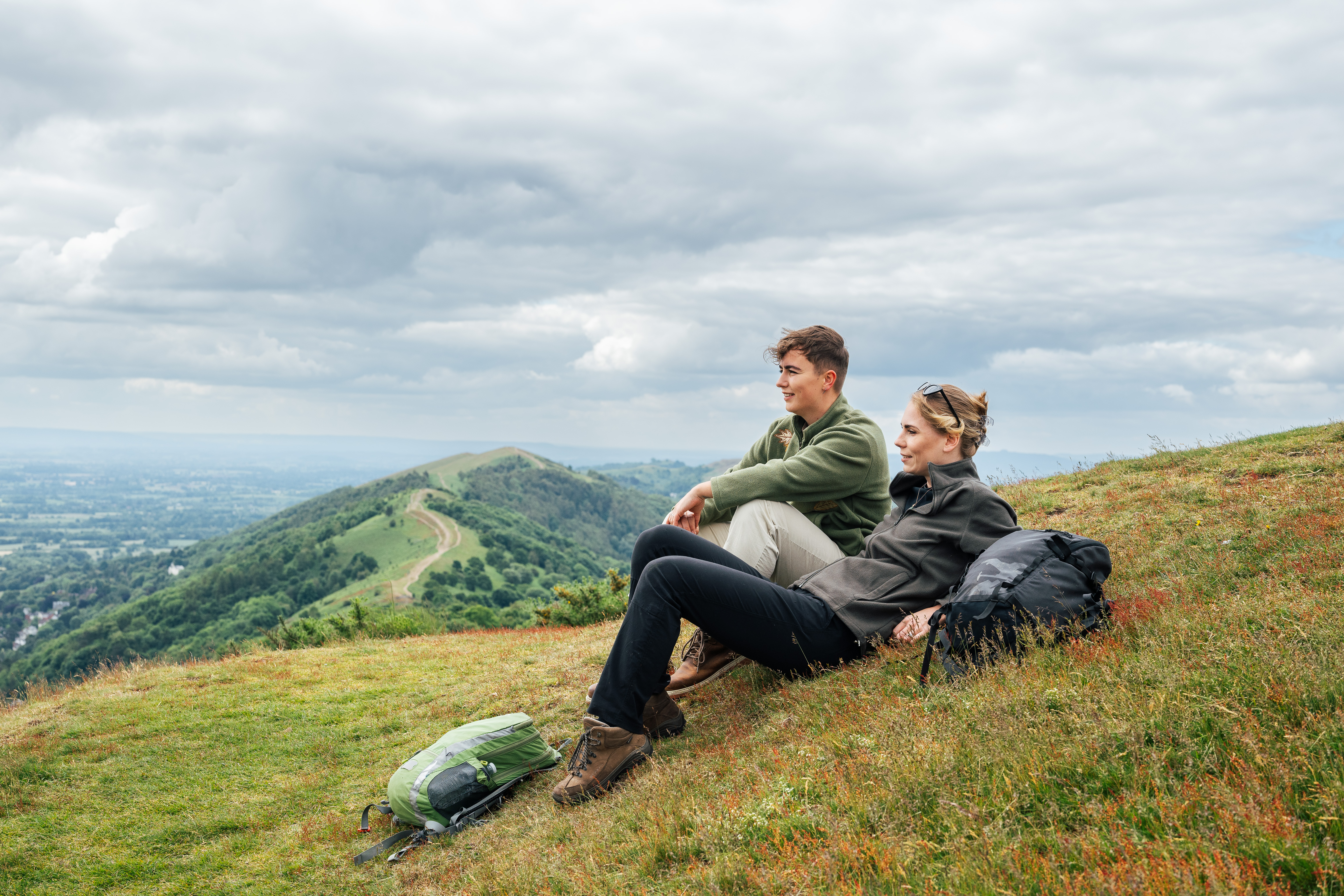 Zwei Frauen sitzen auf einem Hügel und blicken auf eine Aussicht mit grünen Hügeln im Hintergrund