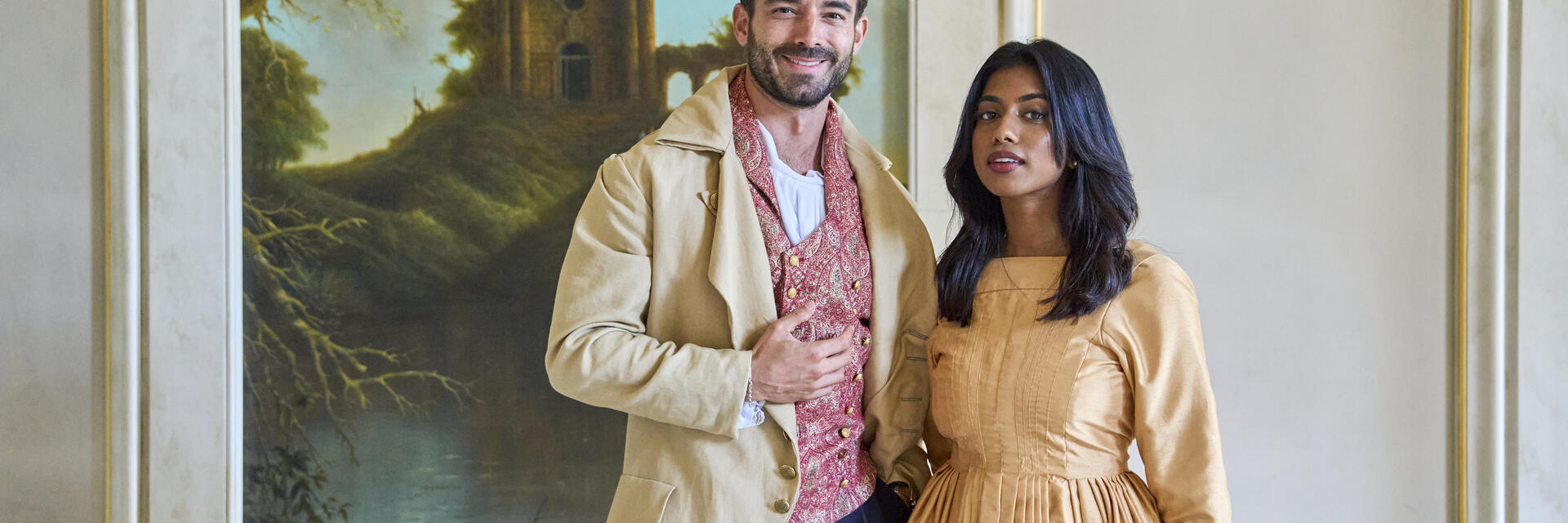 Ein Mann und eine Frau in historischen Kostümen stehen in einem dekorierten Raum vor einem Gemälde mit Landschaft und altem Gebäude.