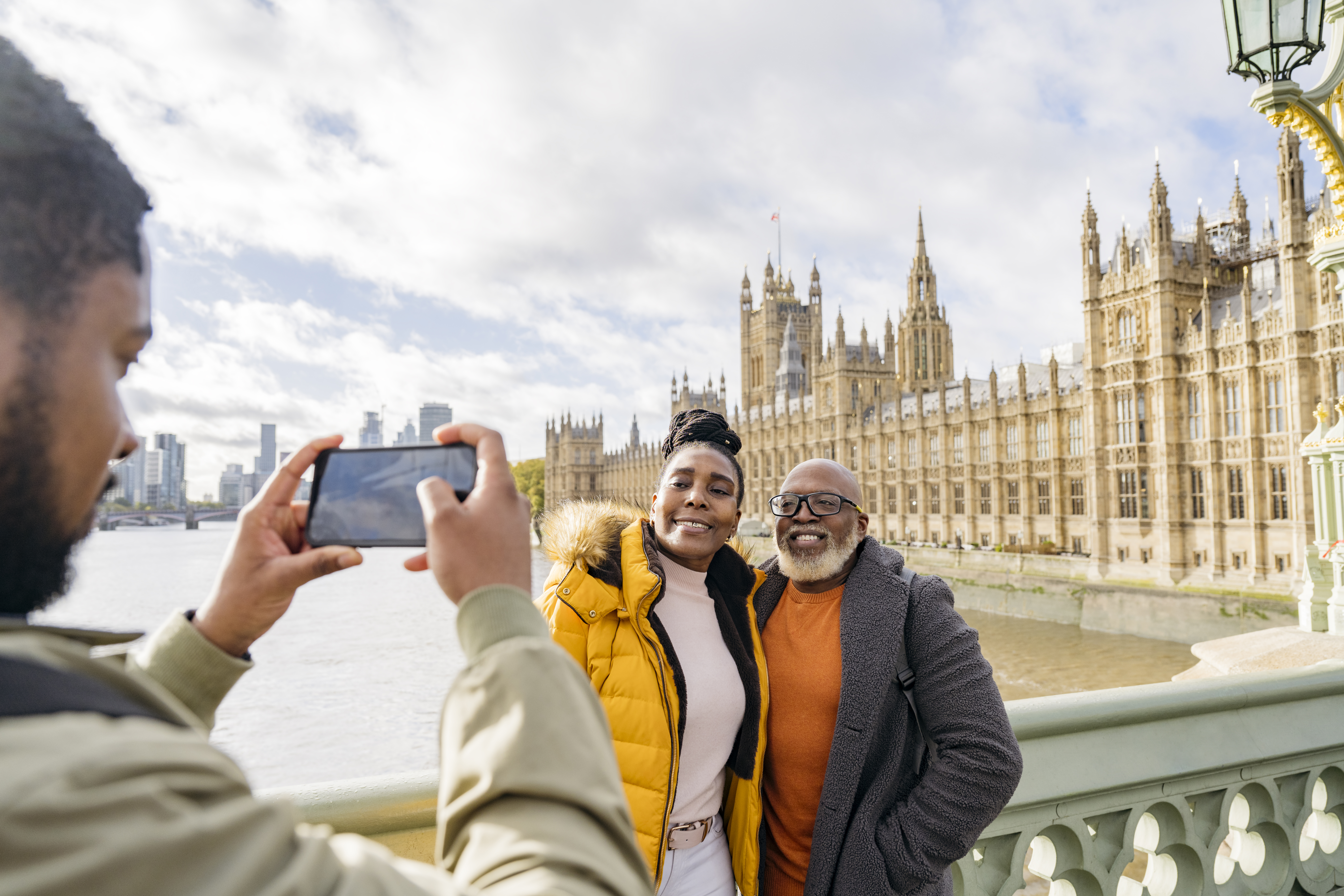 A man photographing mature couple with a large river and iconic buildings in the background.