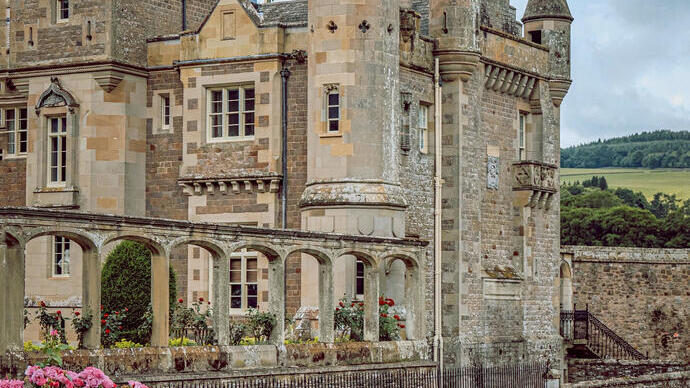 Castillo antiguo con torres, arco de piedra, jardín con flores y mujer con vestido floreado caminando sobre el césped.
