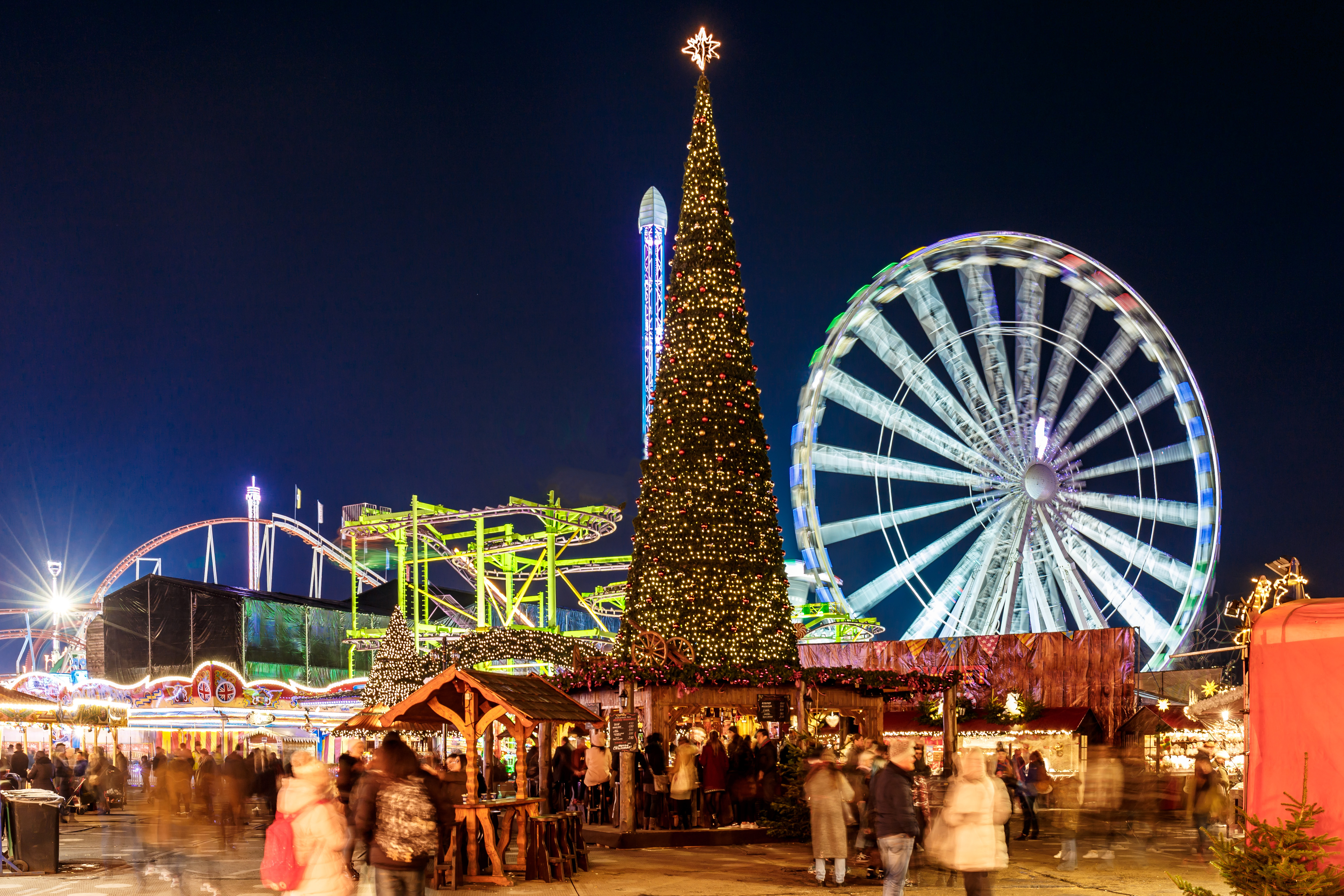 A Christmas fair at night with a large decorated Christmas tree, ferris wheel, roller coaster, lights, and crowds of people.