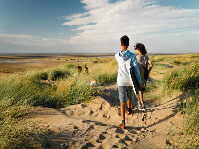 Two people walk with boards along sandy dunes to a wide beach under a blue sky, with two others sitting among the grass.