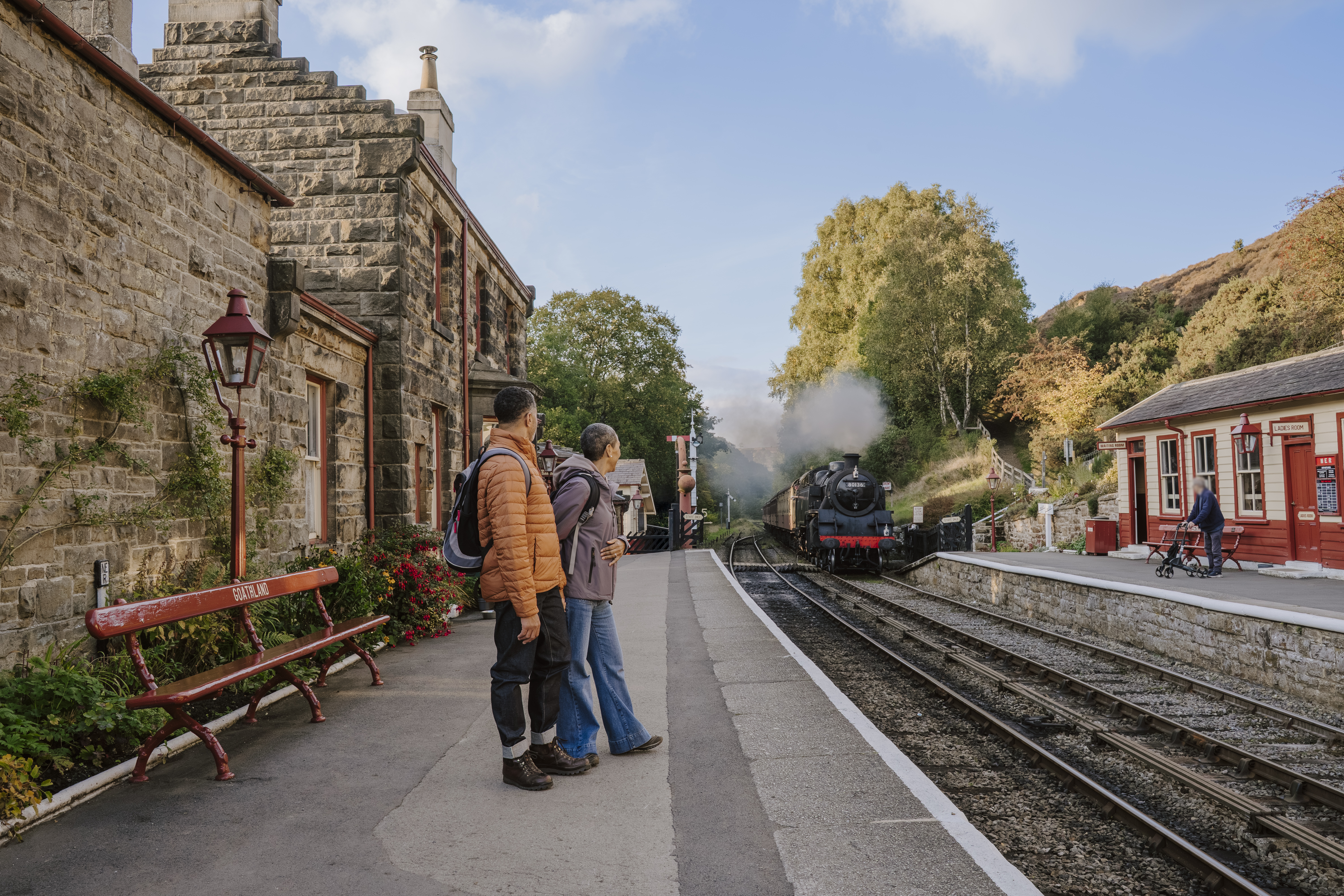 Historic railway station with stone buildings, a steam train arriving, and people waiting on the platform. Autumn trees and clear sky in the background.