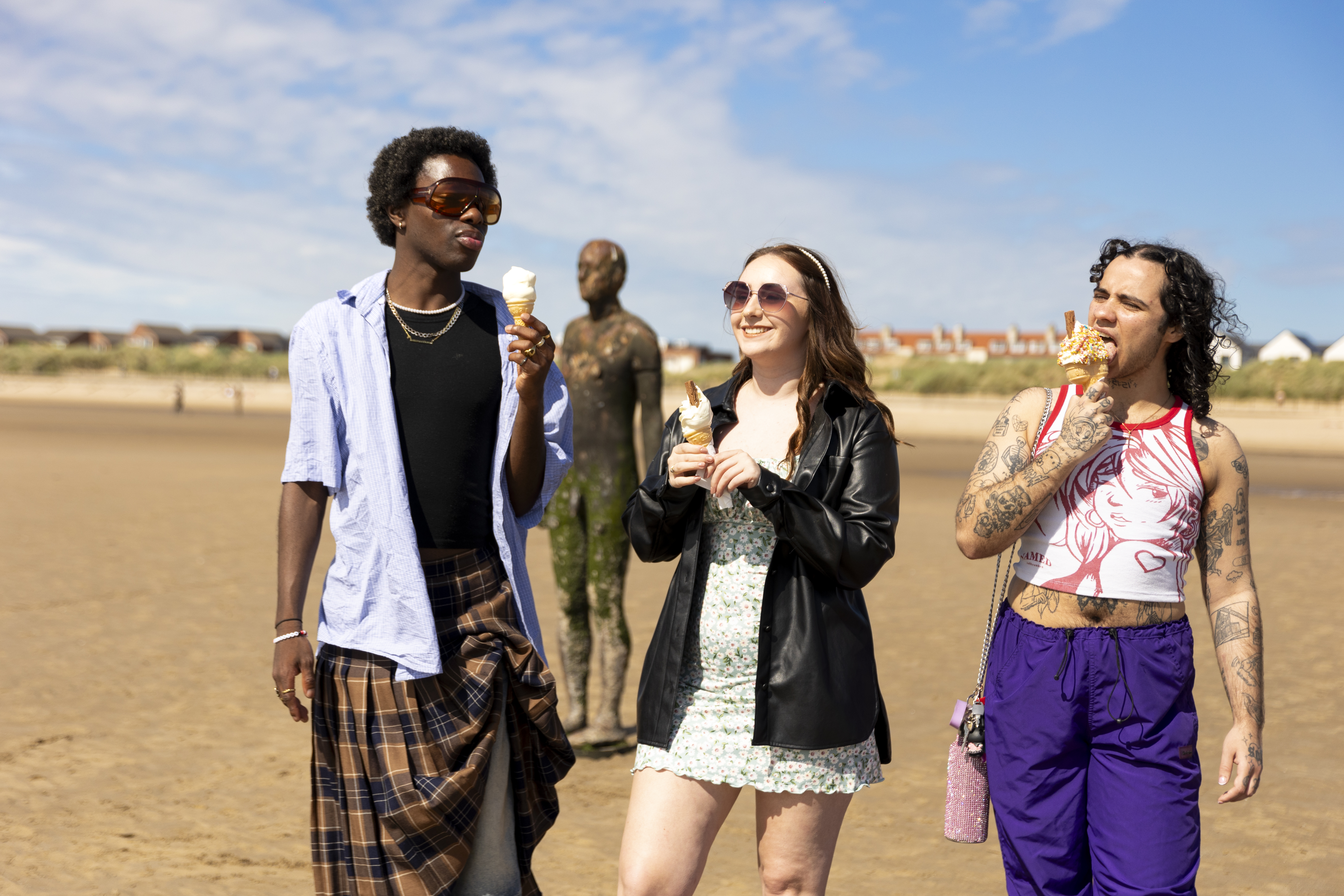 A group of friends walk on the beach with ice creams, a statue in the background