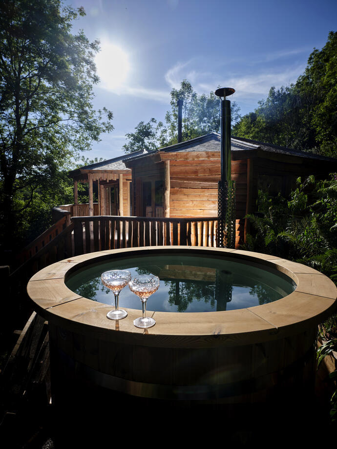 A wooden edged hot tub in front of the lit wooden lodge at Sleepy Owl Devon.
