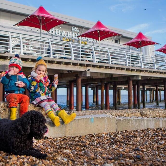 Three children eat ice cream near a pebble beach, with a black dog in front and a pier with red umbrellas in the background.