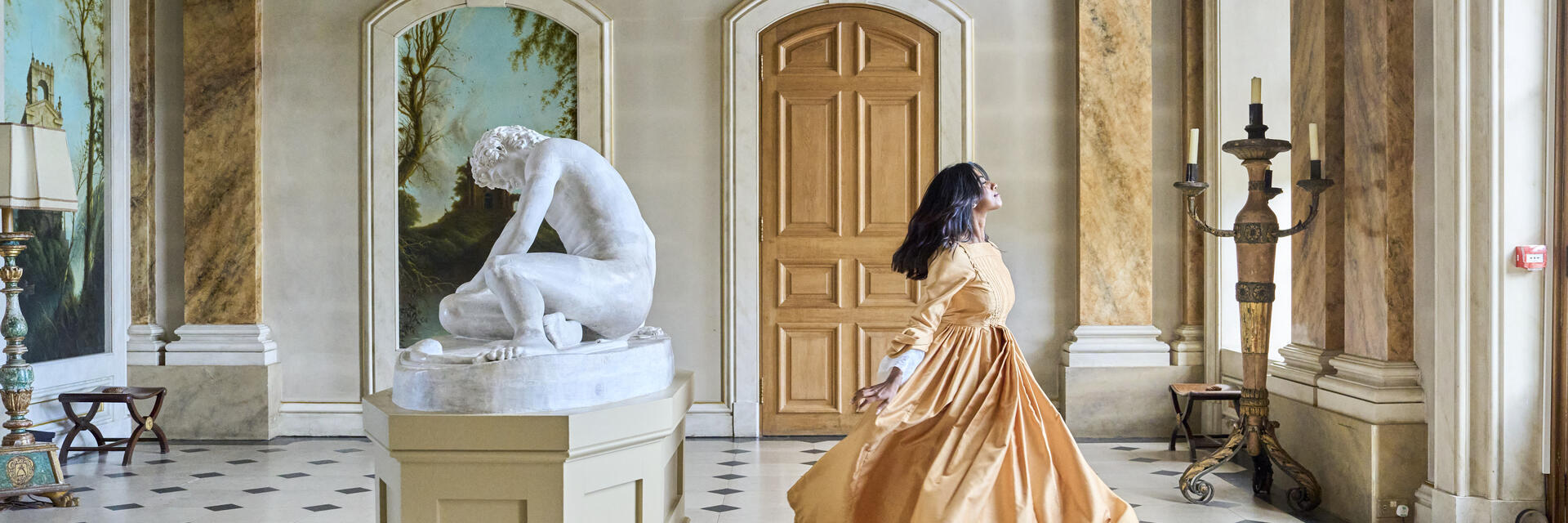 Elegant hall with classical statue, arched doors, large windows, and a person in a flowing period dress spinning gracefully on tiled floor.
