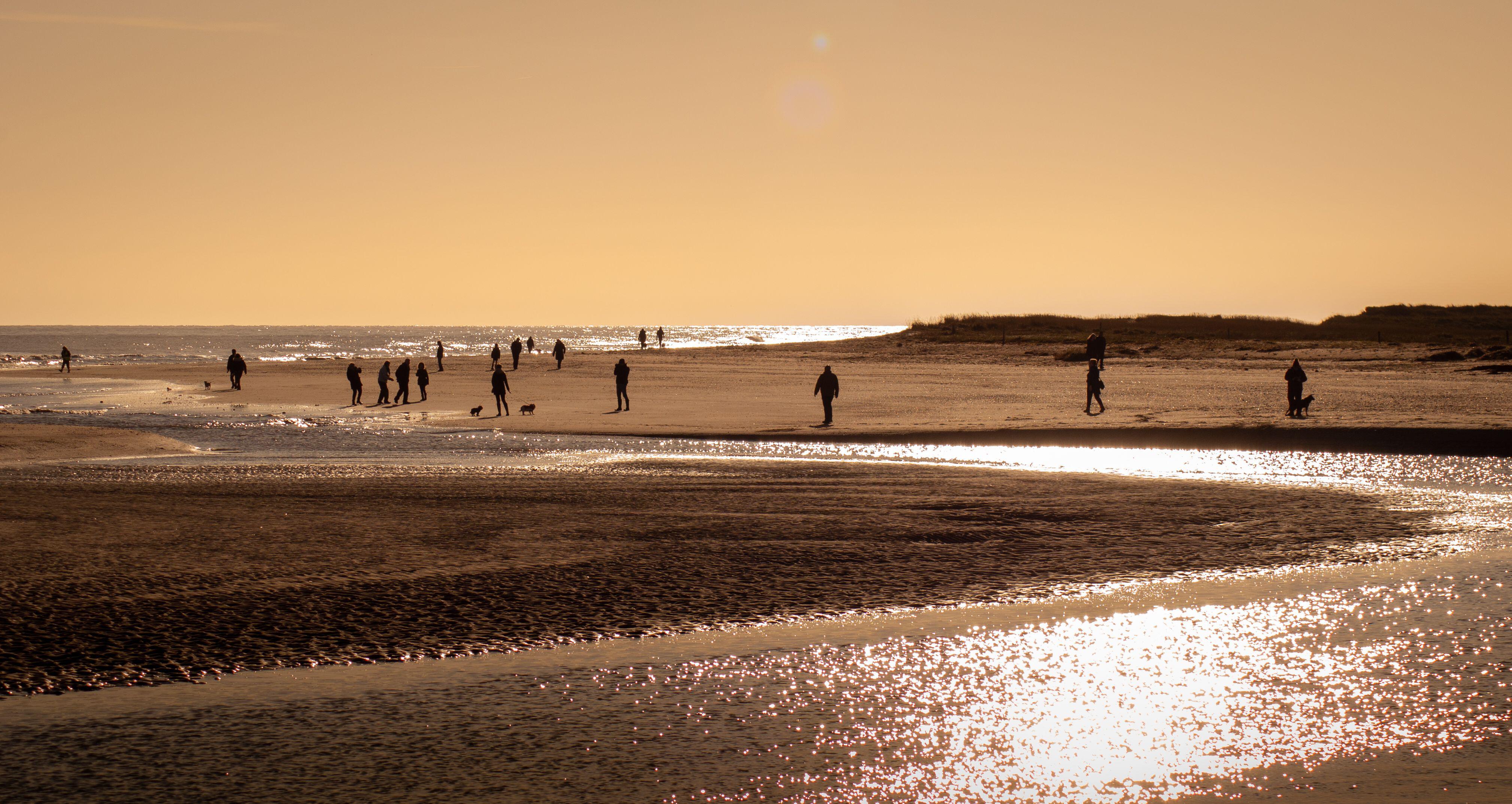 People walk along a sandy beach at sunset, with shimmering water and gentle waves under a warm orange sky.