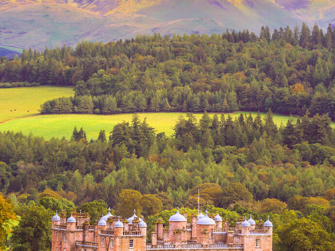 Exterior of a sandstone palace with ornate turrets surrounded by woodland with high mountains in the distance in autumn.