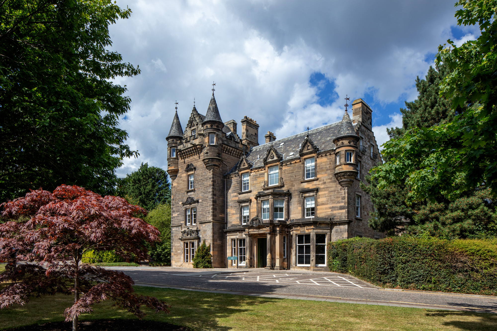 Historic stone castle-like mansion with turrets, surrounded by trees and a manicured lawn under a partly cloudy sky.