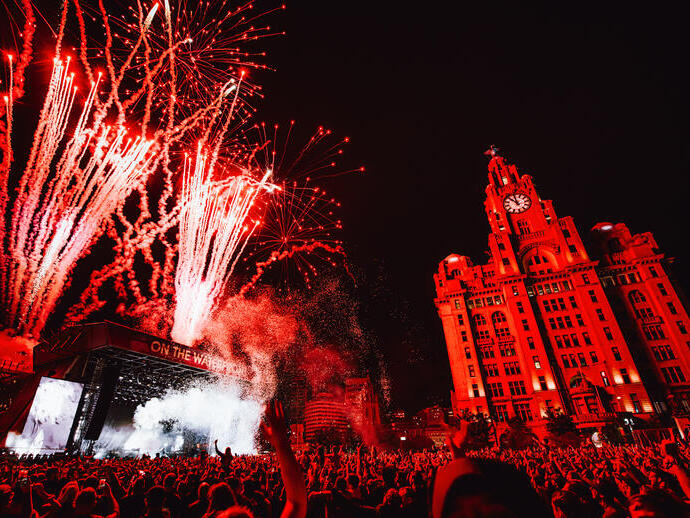 Crowds of people watching a band at a music festival as fireworks explode, in front of the Liver Building.