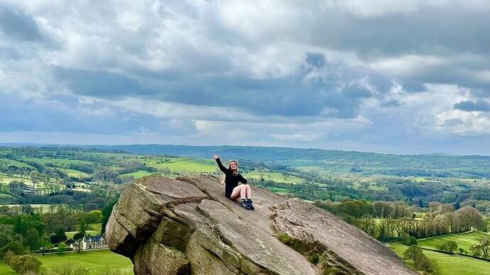 Person sits on large rocky outcrop with scenic countryside and dramatic cloudy sky in the background.