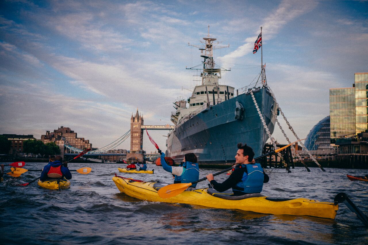 People in Kayaks on a river past iconic navy ships and iconic city landmarks.