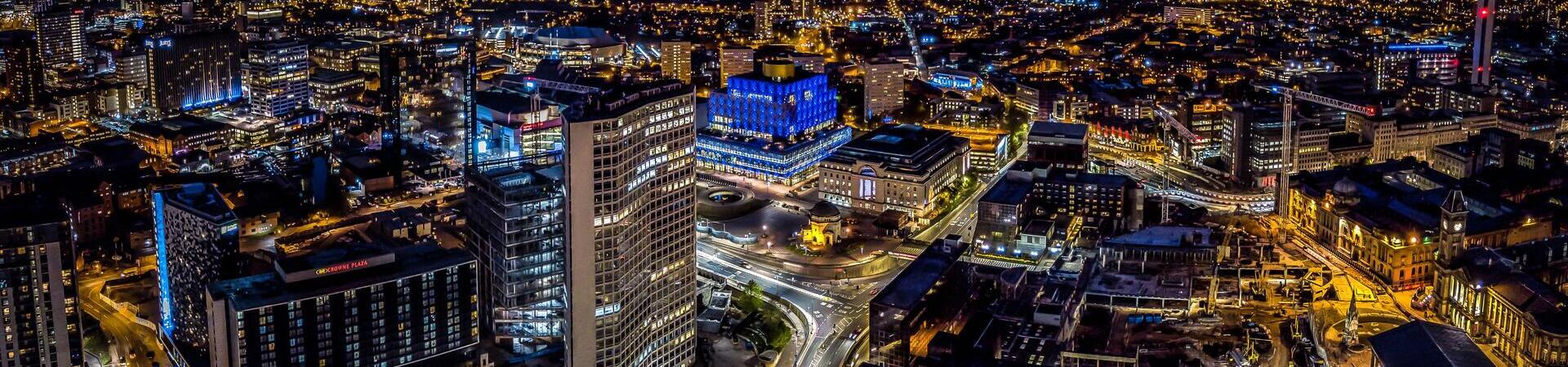 Aerial view of a modern city skyline at dusk, with high-rise buildings, illuminated streets, and a glowing blue-lit structure.