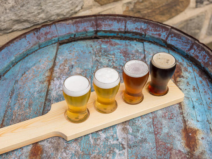 Quatre types de bière dans des petits verres sur une planche en bois, posés sur un tonneau bleu vieilli avec un mur en pierre en arrière-plan.