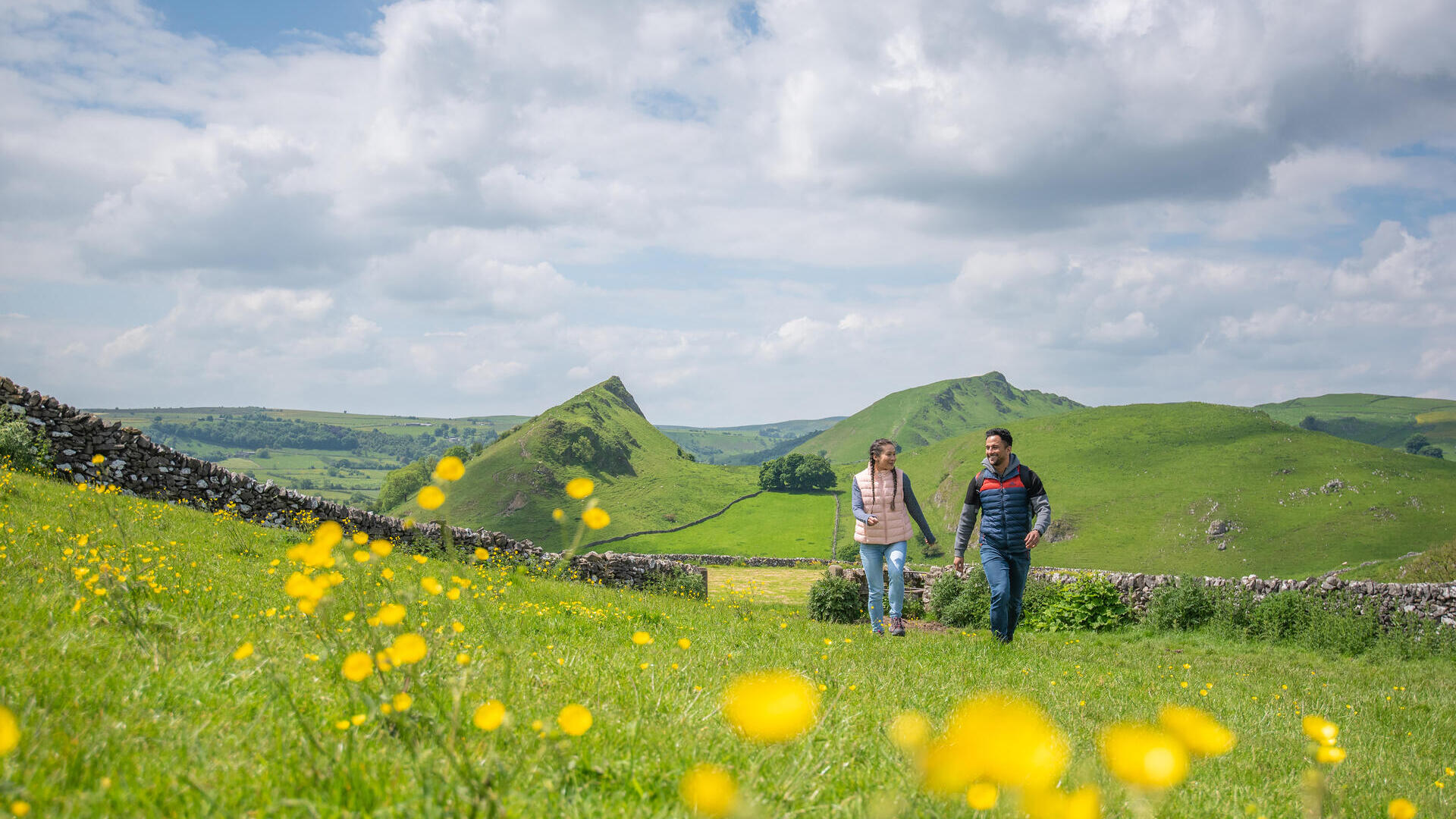 شخصان يسيران في مرج أخضر مزهر مع تلال في الخلفية تحت سماء غائمة.