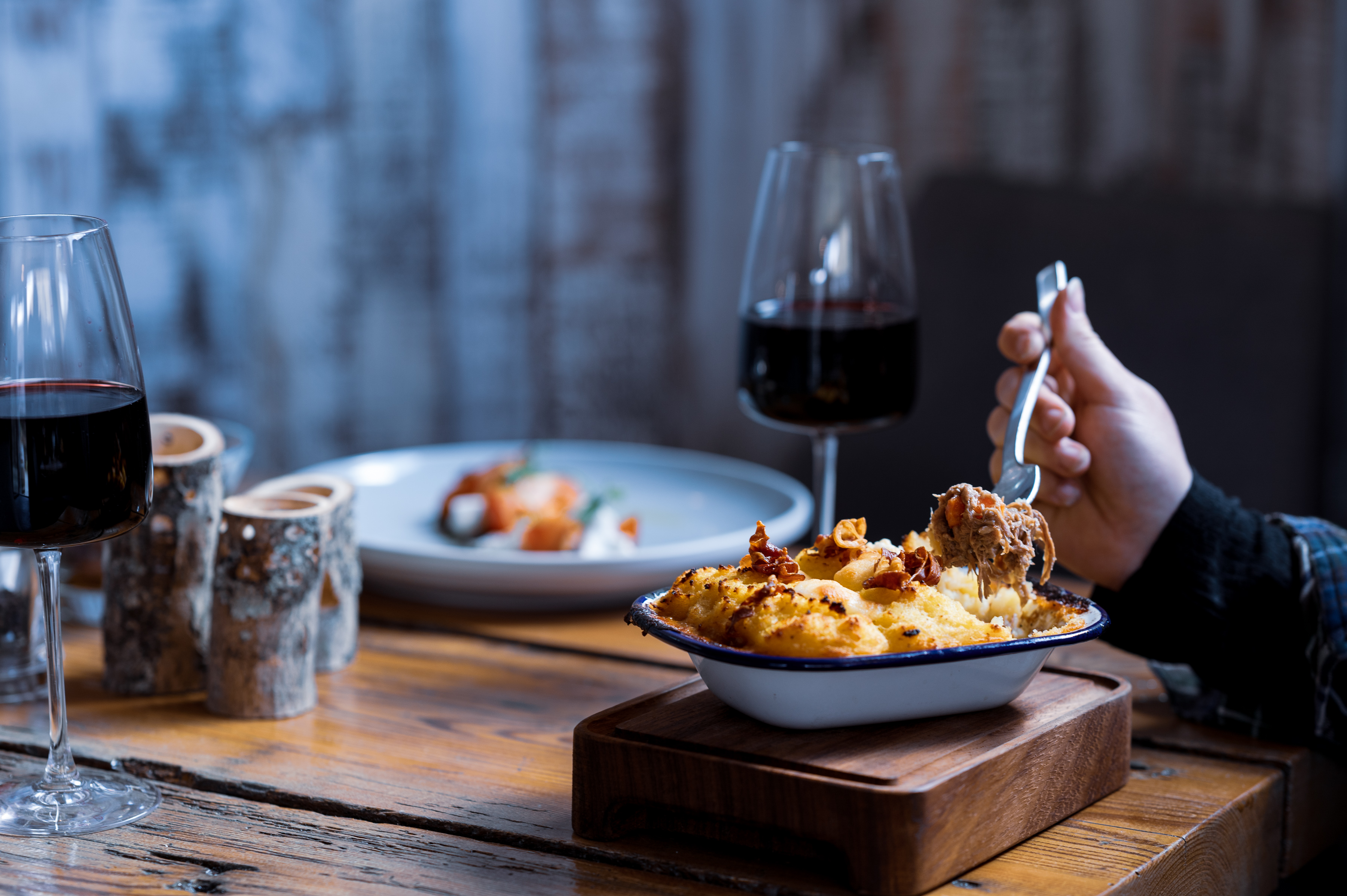 View of arm eating a mashed potatoes topped dish with two glasses of red wine and another dish of food in soft focus in the background 