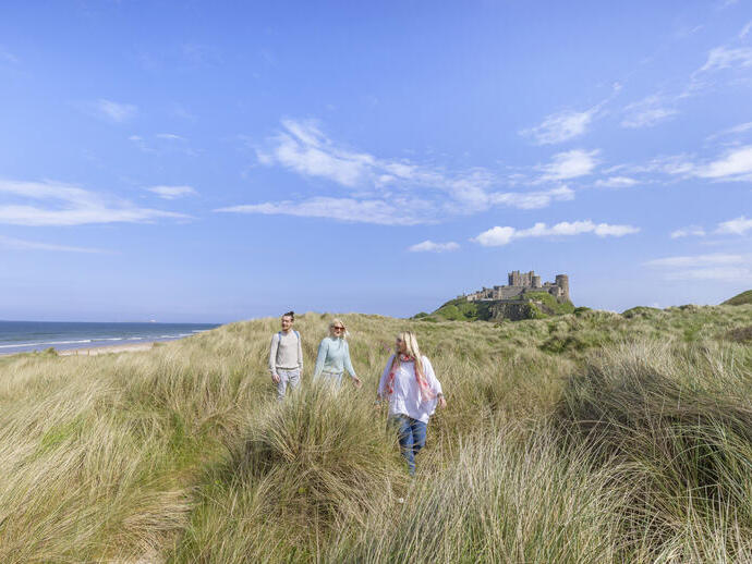 A group of people walk among tall grasses in dunes with a castle in the background
