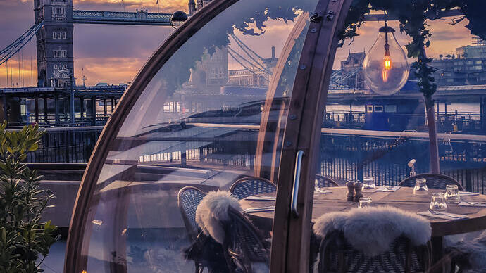 A glass dome enclosing a restaurant table beside a river in the middle of a city