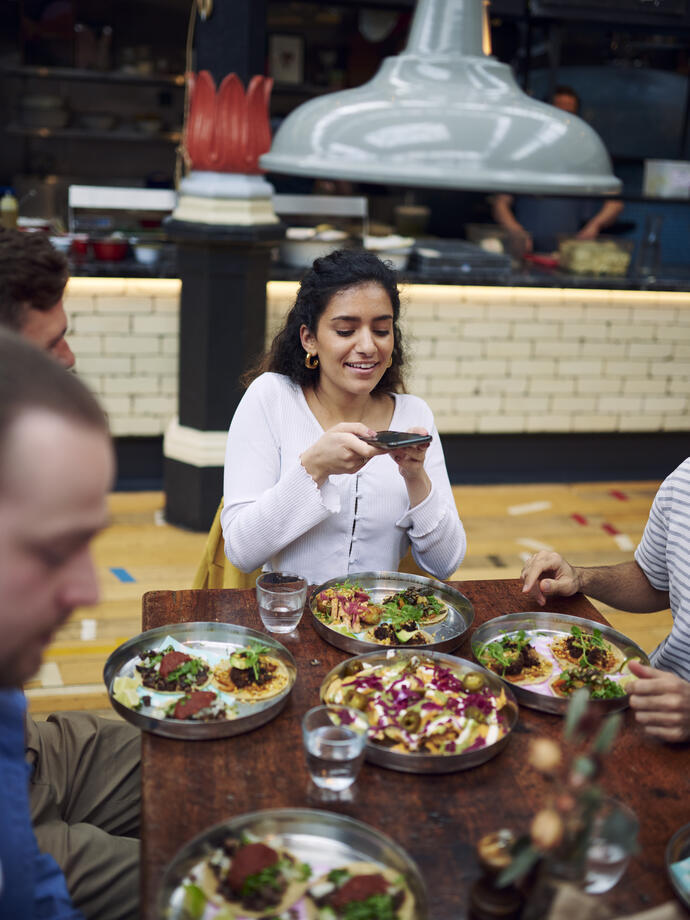 Femme assise avec des amis à une table, prenant une photo des plats dans une aire de restauration