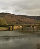 A stone and rusted metal bridge across a wide river in the countryside in autumn.