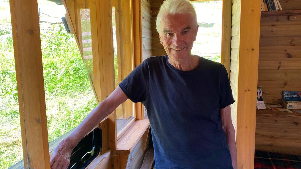 Person standing inside a wooden cabin with open windows, bookshelves, and greenery visible outside.