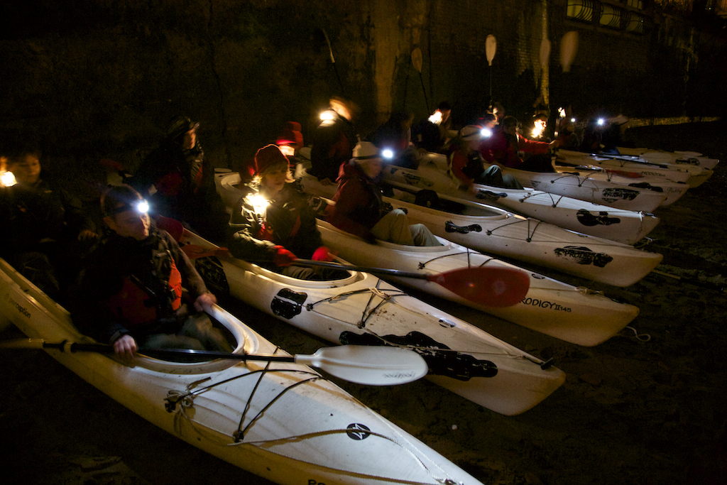 Row of people with head torches sat waiting in white canoes in water ready to go out for the night tour