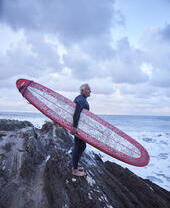 Man carrying a surfboard, standing on the rocks looking out to sea