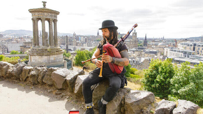 Mann spielt Dudelsack, während er auf einem Felsen auf einem Hügel sitzt und auf eine Stadt blickt