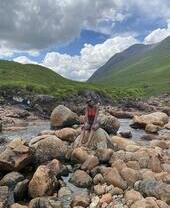Frau sitzt auf einem Felsen in einem Bach, umgeben von Bergen
