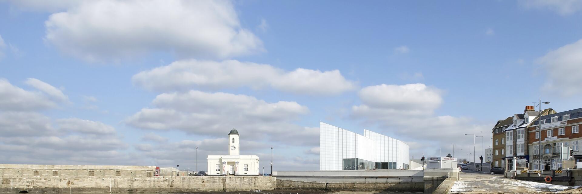Modern building overlooking a harbour and beach, a row of townhouses in the background