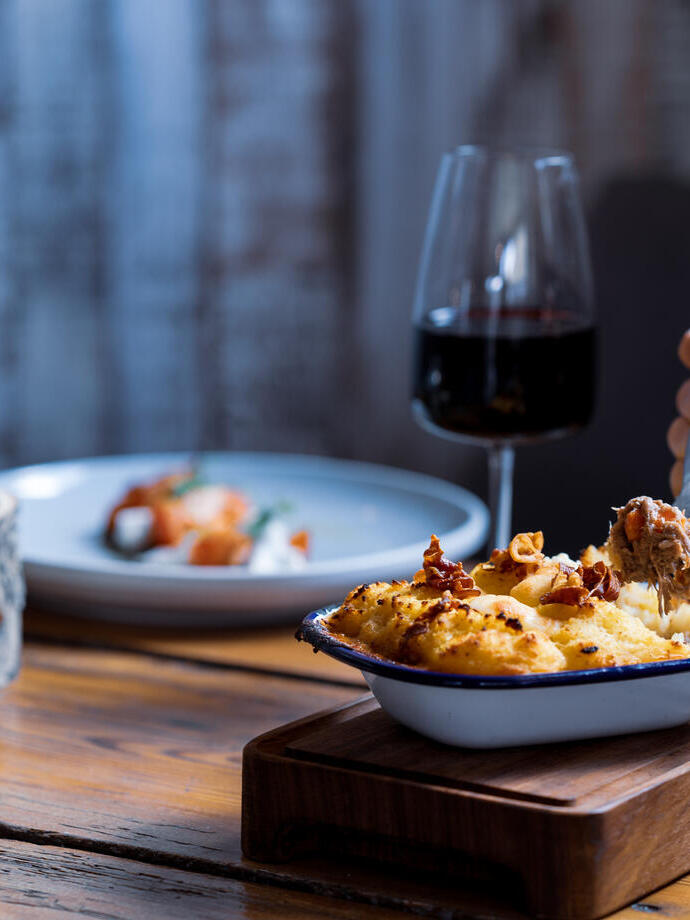 View of arm eating a mashed potatoes topped dish with two glasses of red wine and another dish of food in soft focus in the background 