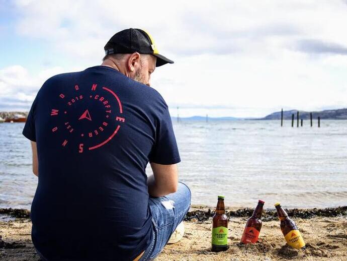 Ein Mann sitzt am Strand neben Flaschen Snowdon Craft Beer