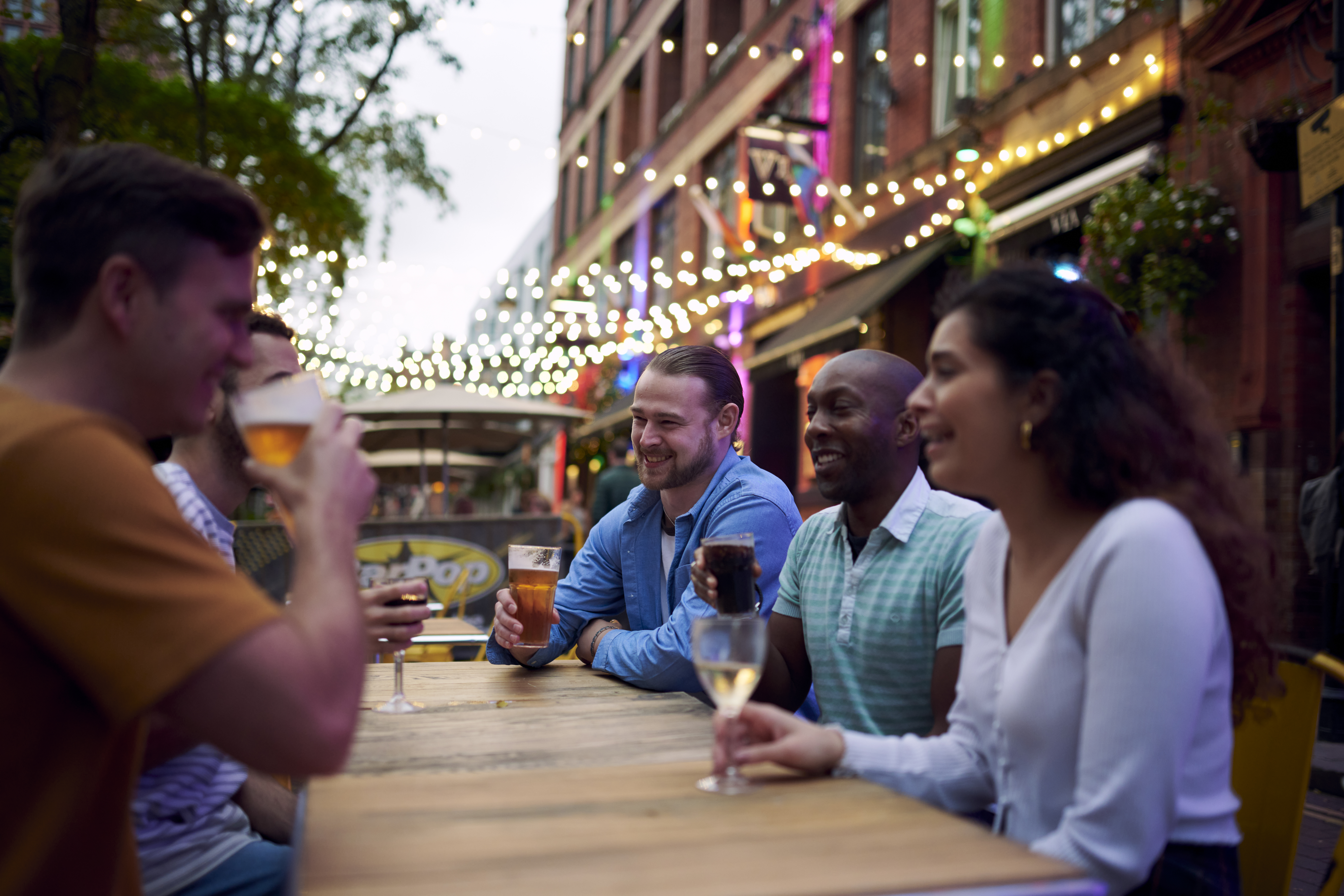 Friends having a drink sat at an outside table of a bar with fairy lights hanging behind