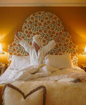 Person in white robe relaxing on a bed in a warmly lit, stylish bedroom with patterned headboard and yellow walls.