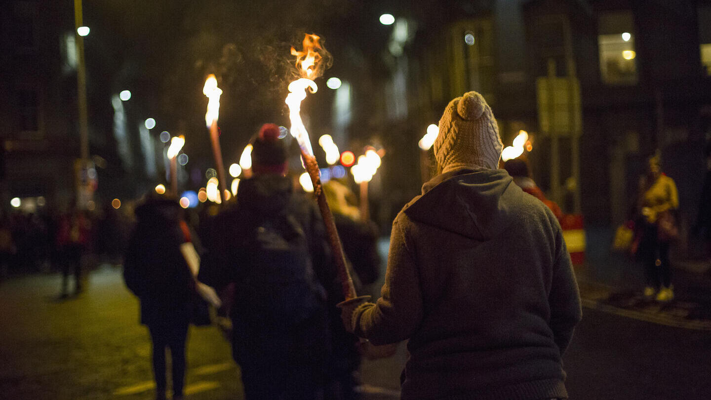People marching at night on a city street holding flaming torches, with urban buildings and streetlights in the background.
