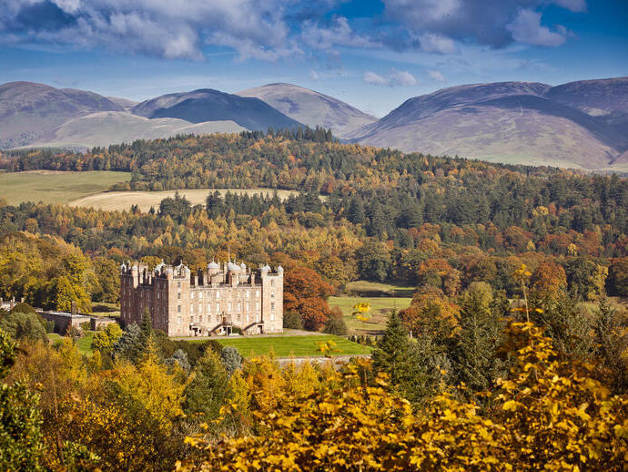 A historic castle set amid vibrant autumn trees, green fields, and hills under a bright blue sky.