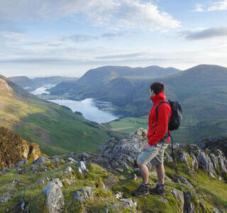 A man wearing a rucksack stands on a mountain fell overlooking hills and a lake