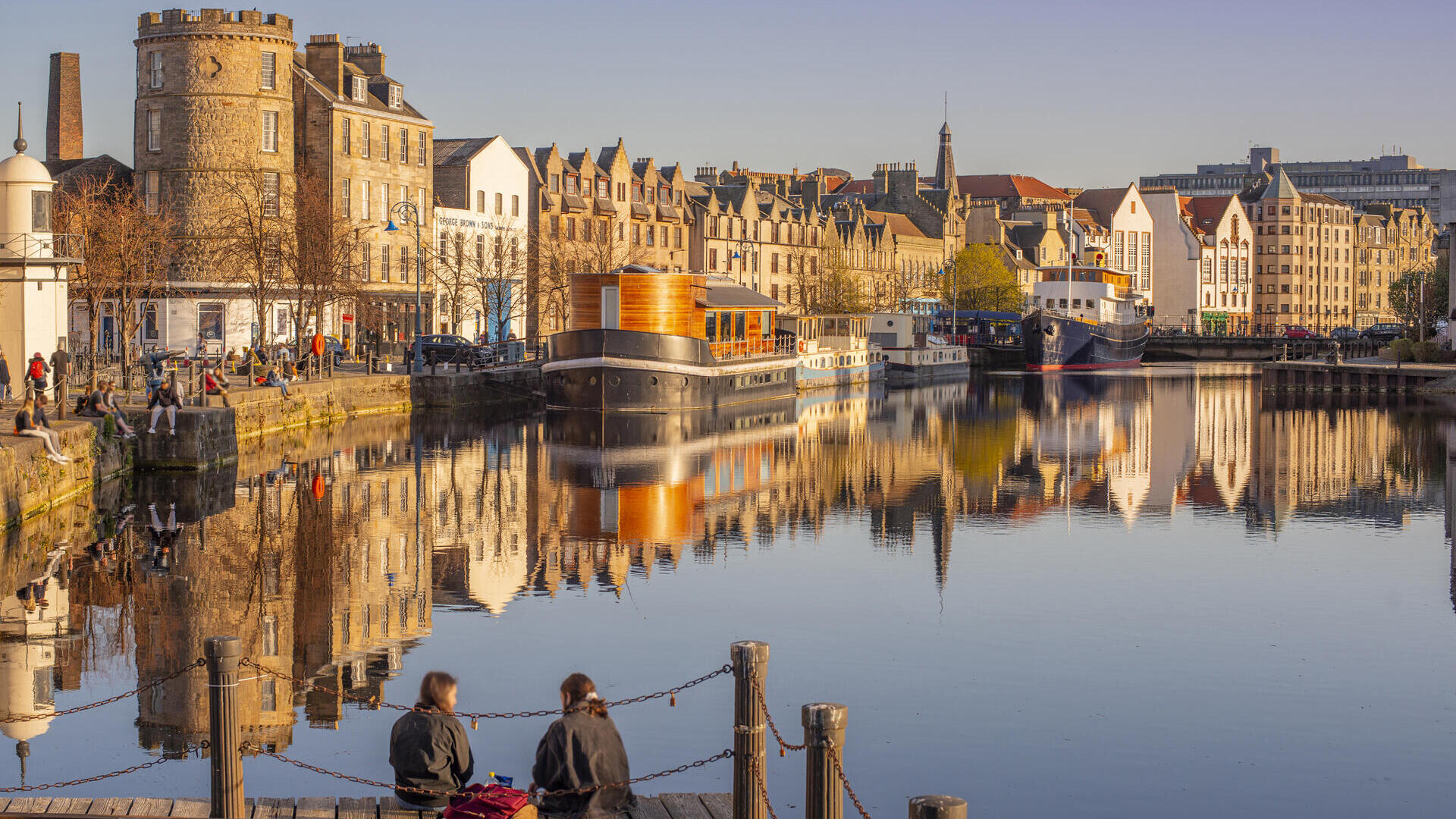 Two young women sat on the jetty of the waterfront in the evening with boats and buildings by the harbour.