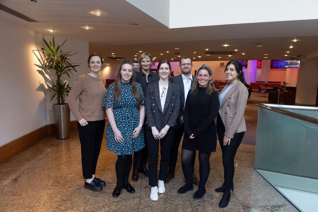Six women and one man in professional clothing stand together posing in the hallway of a large convention centre.